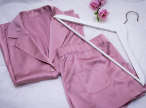 A set of pink silk pajamas laid out on a bed next to a clothing hanger and a flower