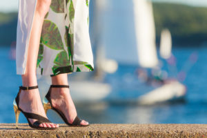 Woman wearing high heeled sandals, standing in front of a body of water with sailboats on it