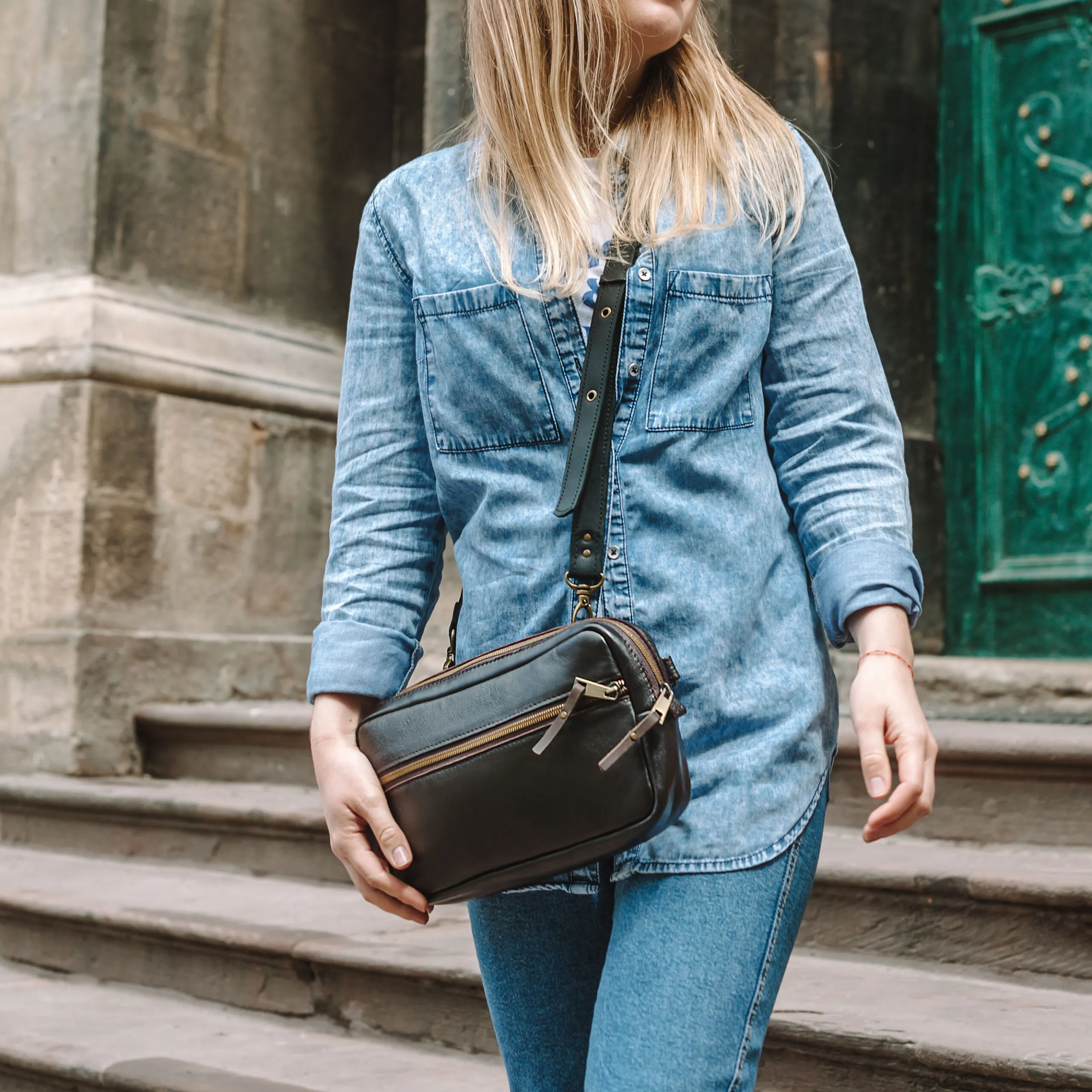Leather sling bag for women. Belt Bag Minimalist. Blond woman in denim shirt and, blue jeans and a dark brown leather bag over the shoulder. Close-up.