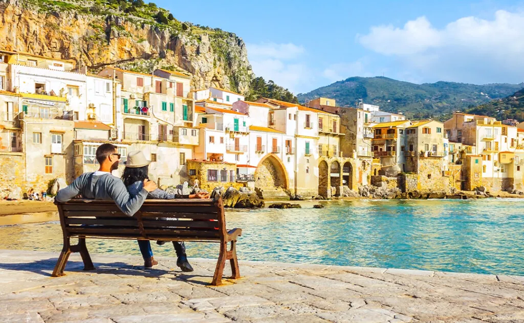 Couple sitting on bench, enjoying view of beach town of Cefalu in Sicily, Italy
